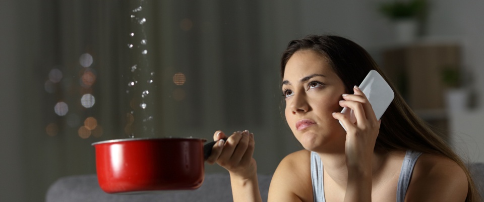 Frustrated woman talking on the phone and holding a pot because water is dripping from the ceiling to illustrate when to submit a maintenance request.