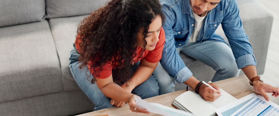 Two renters sitting on a couch reviewing bills and writing in a notebook, planning expenses and learning how to budget for an apartment together.