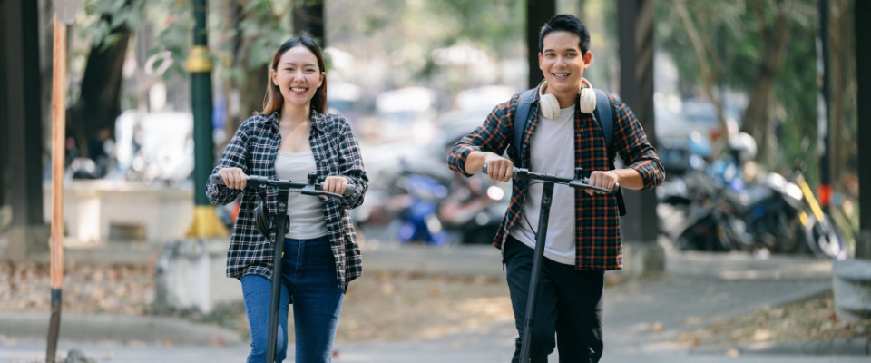 Two students smiling while riding electric scooters through a park on a sunny day, representing modern public transit and sustainable urban mobility.