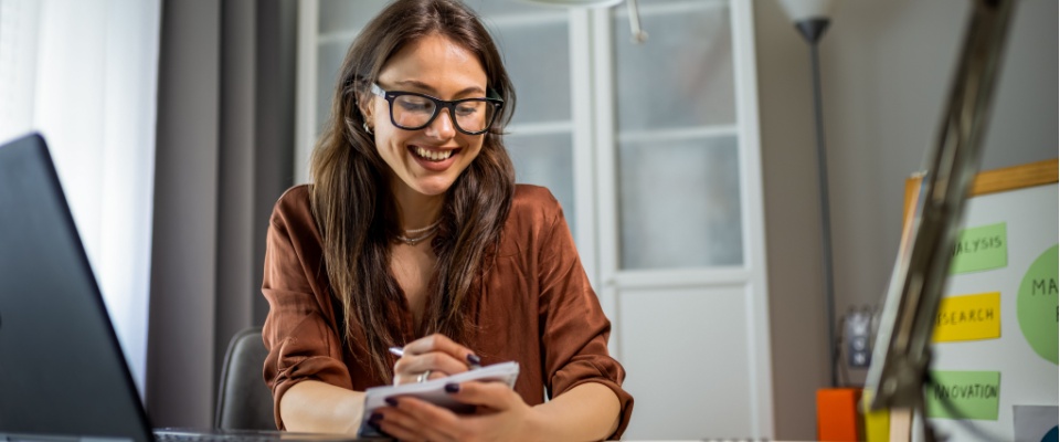 Focused young woman wearing glasses works from home with her laptop, writing notes on paper for better organization and productivity to illustrate a successful lease renewal outside of peak season.