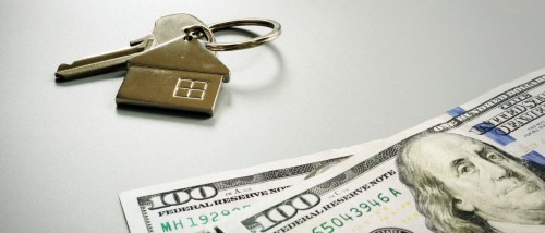 House-shaped keychain and apartment keys placed next to U.S. dollar bills, symbolizing rent collection, monthly rent payments and the cost of renting an apartment.