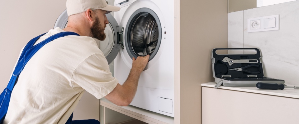 Maintenance worker inspecting a dryer in a shared laundry room with tools nearby, highlighting the importance of reporting broken machines promptly.
