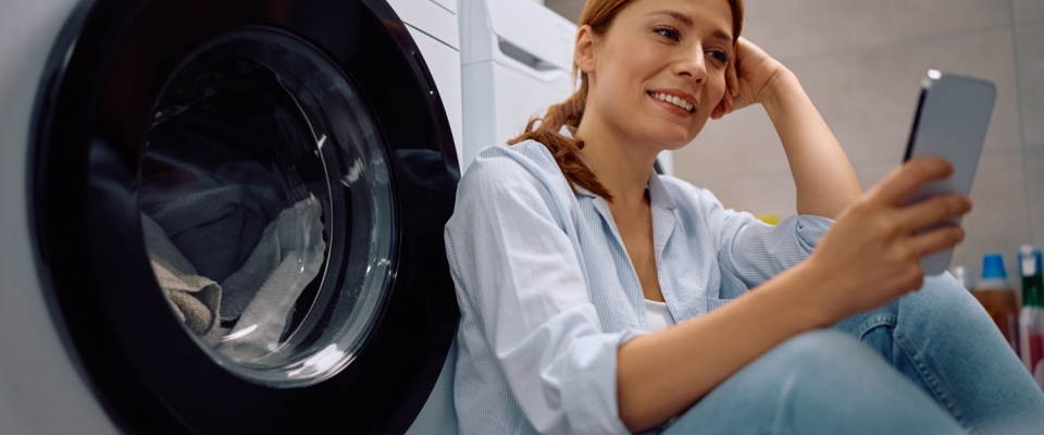 Resident sitting on the floor of a shared laundry room next to a front-loading washer, checking her phone to set a timer while waiting for her laundry cycle to finish.