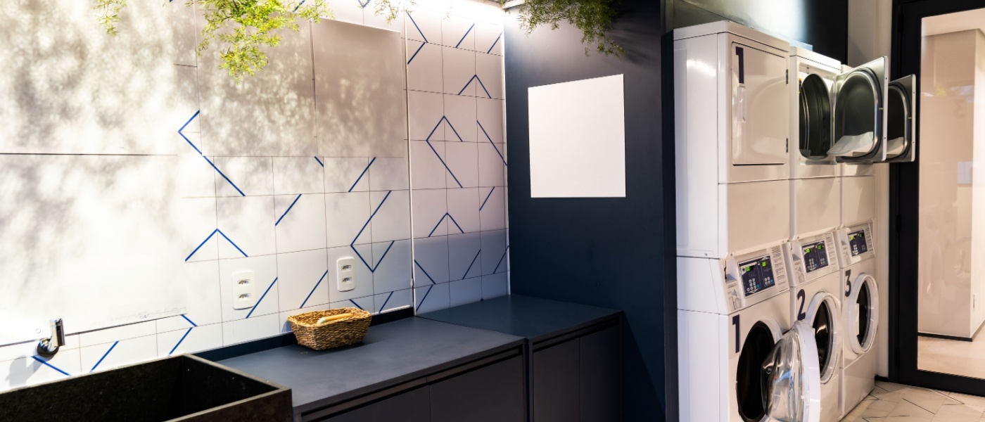 Modern shared laundry room in an apartment building with stacked washers and dryers, folding counter space, tiled walls and indoor plants for decor.
