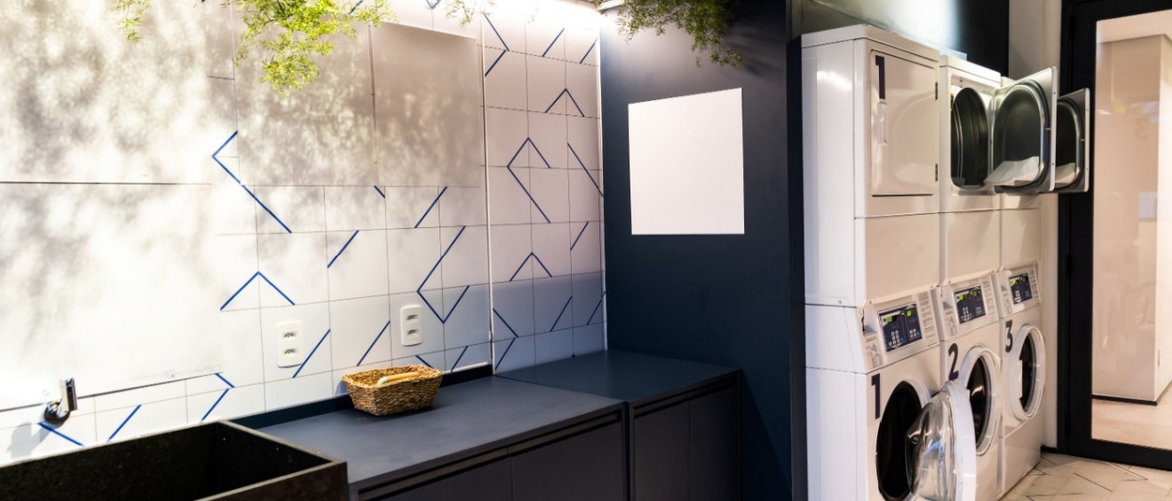 Modern shared laundry room in an apartment building with stacked washers and dryers, folding counter space, tiled walls and indoor plants for decor.