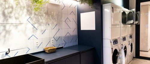 Modern shared laundry room in an apartment building with stacked washers and dryers, folding counter space, tiled walls and indoor plants for decor.