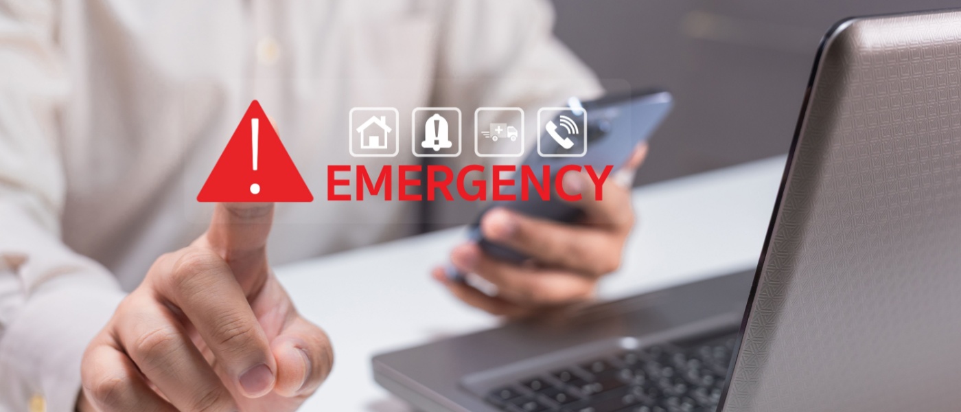 A person is alerted to an emergency situation, using a smartphone while pointing at a laptop with an "EMERGENCY" notification displayed prominently to illustrate an emergency contact.