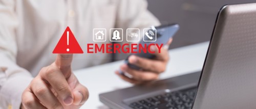 A person is alerted to an emergency situation, using a smartphone while pointing at a laptop with an "EMERGENCY" notification displayed prominently to illustrate an emergency contact.