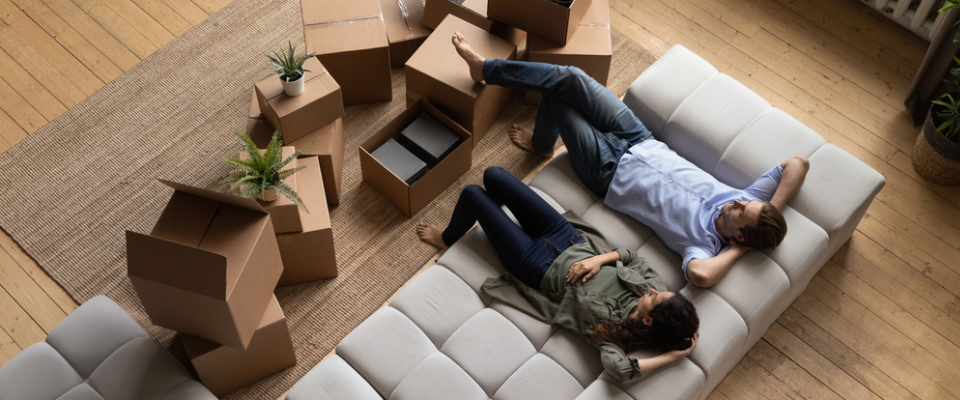 Renters relaxing on a sofa surrounded by packed boxes in a bright apartment, symbolizing moving to a new place and navigating the cost of living.