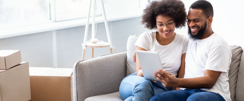 Couple sitting on a sofa using a tablet to search for an apartment online, reviewing a rental application together as co-applicant partners.