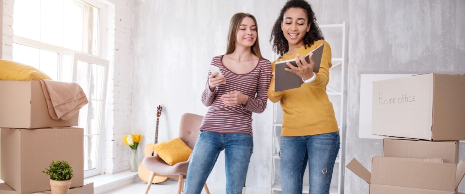 Two young women holding a smartphone and an agenda in an apartment filled with boxes to illustrate an apartment cosigner and a coapplicant.