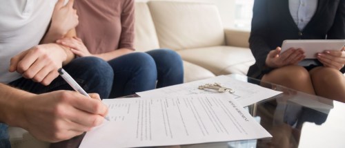 A closeup of several people around a coffee table with someone signing the rental agreement to illustrate an apartment cosigner versus a coapplicant.