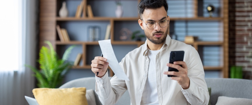 Young man sitting on a sofa at home, holding a receipt and checking his phone with a concerned expression, reviewing expenses and a rent receipt.
