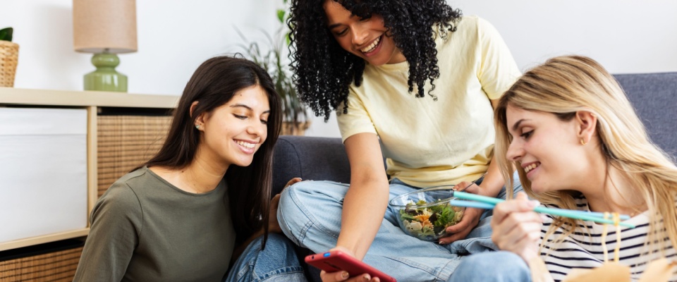Young roommates relaxing on a sofa in their shared apartment while using a mobile phone, representing shared living and financial responsibilities like a security deposit.