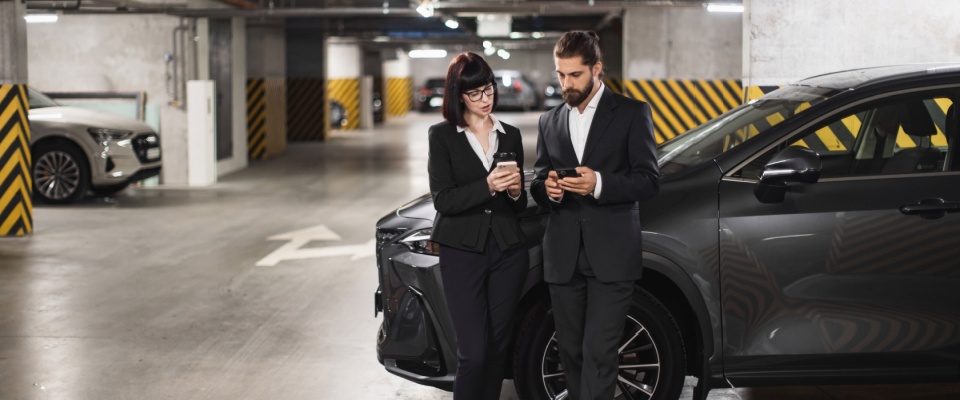A man and a woman standing by a car in a garage and looking at their smartphones to illustrate neighbors discussing tandem parking issues.