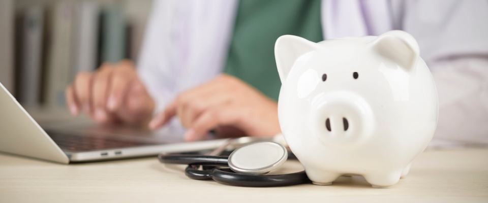 White piggy bank with a stethoscope on a desk and a doctor working on a laptop in the background, symbolizing financial planning and savings similar to setting aside money for a security deposit.