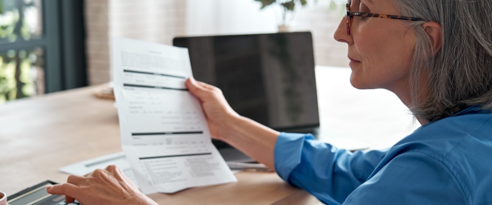 Senior businesswoman sitting at a desk at home, holding a bill and using a calculator to manage finances, illustrating budgeting and expenses like a rental application fee.