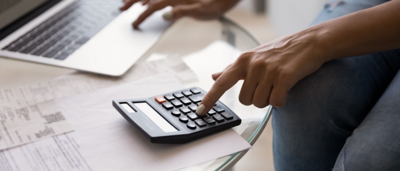 Young woman using a calculator and online banking on her computer to manage monthly expenses, illustrating budgeting and costs like a rental application fee.