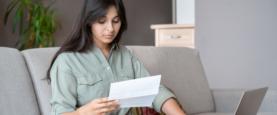Young woman using a calculator and online banking on her computer to manage monthly expenses, illustrating budgeting and costs like a rental application fee.