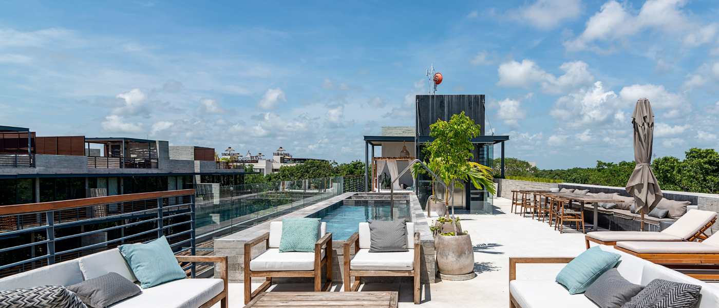 Rooftop community amenities featuring a lap pool, lounge seating with teal and gray cushions, sun loungers, a high-top bar area, and lush greenery in the background under a bright blue sky.