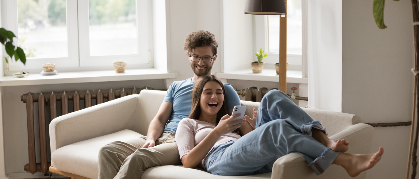Happy couple relaxing on a couch in their apartment while browsing flexible rent payment options on a smartphone.