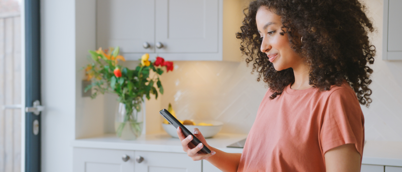 Smiling renter in her apartment kitchen using a smartphone to set up rent payment reporting and build her credit score.