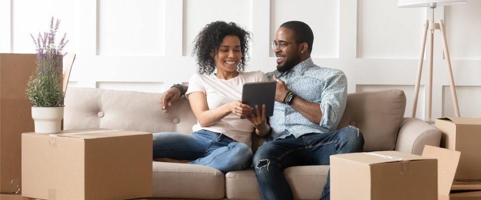 Happy couple sitting with moving boxes in a new home while using a tablet, representing relocation planning and a cost of living comparison between cities or neighborhoods.