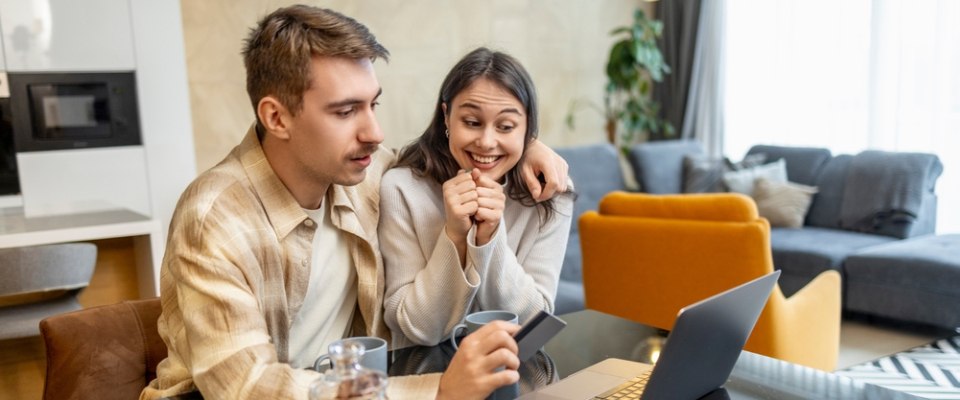 Young couple sitting on a couch, smiling as they look at a laptop and hold a card, comparing the cost of living in two cities.