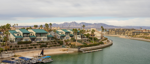 Waterfront homes with green roofs and palm trees line a calm channel in Lake Havasu City, AZ, a growing destination for millionaire renters, with boats docked in the foreground and mountains in the distance.