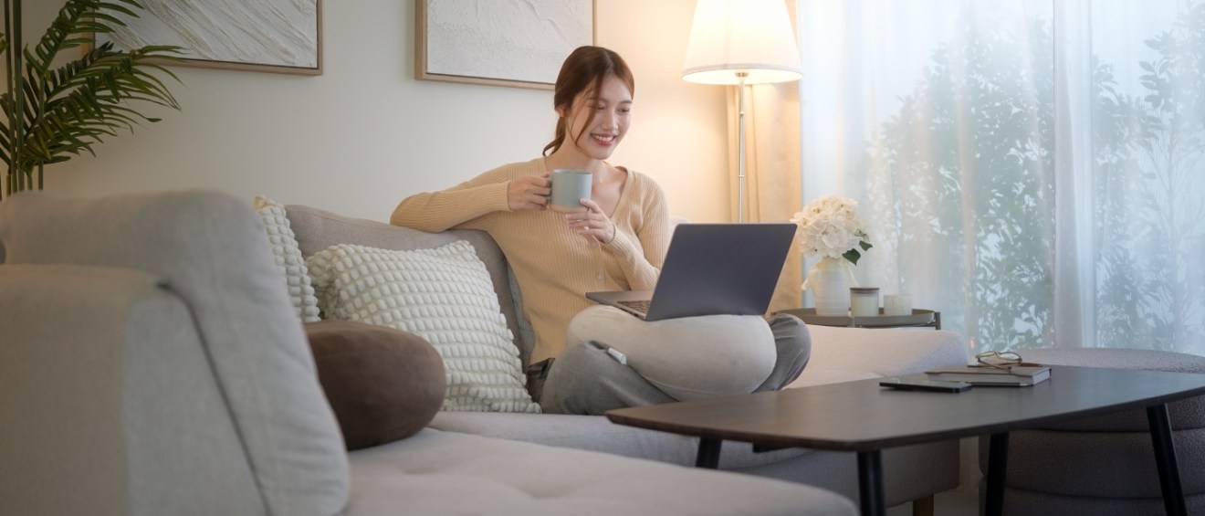 Young woman relaxing on a sofa with a laptop and a hot drink in a cozy living room, illustrating the comfort and independence of solo apartment life.