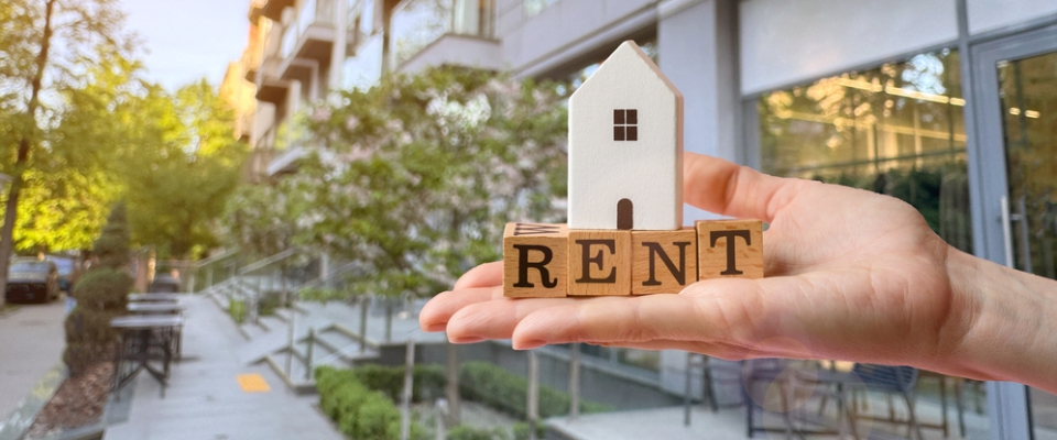 Hand holding a small white model house above wooden blocks spelling RENT, with a modern apartment building behind.