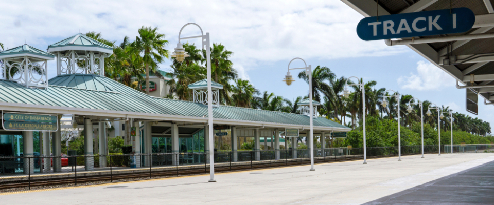 Tri-Rail platform under a green-roofed shelter with palm trees, a public transit option connecting Boca Raton to the wider South Florida region.