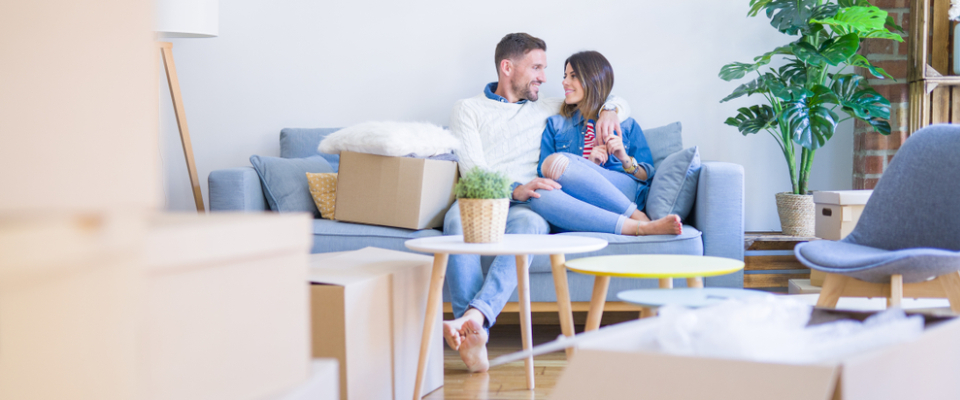 Relaxed couple on a blue sofa amid moving boxes in a bright apartment, having considered the cost of living before their move.