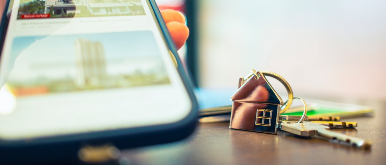Person searching for apartments on a phone next to a house-shaped keychain and keys on a desk.