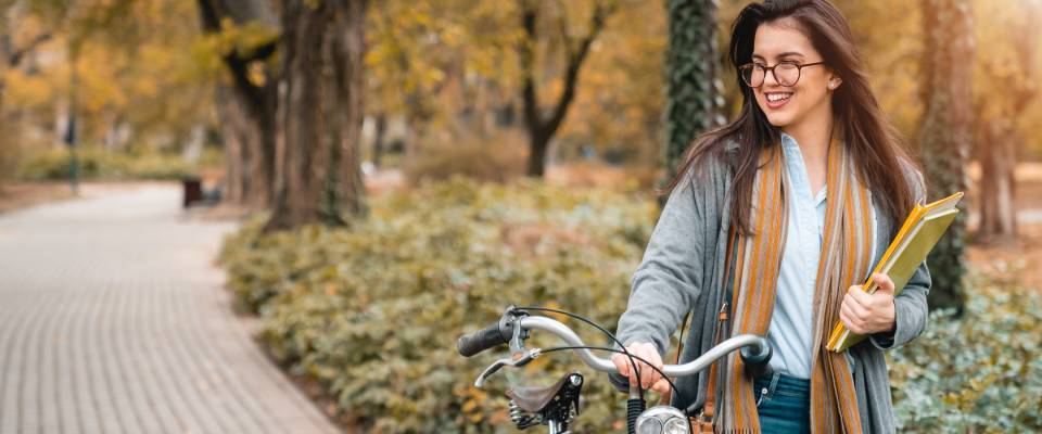 Young smiling female student holding books while walking with a bike in a public park in one of the walkable cities, beautiful girl posing with bike outdoors.