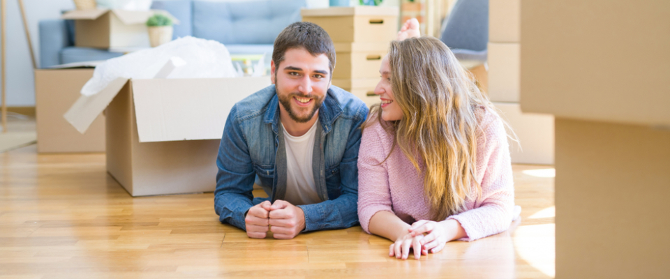 Young couple lying on the floor of a new apartment surrounded by moving boxes, illustrating a cost of living comparison for renters.