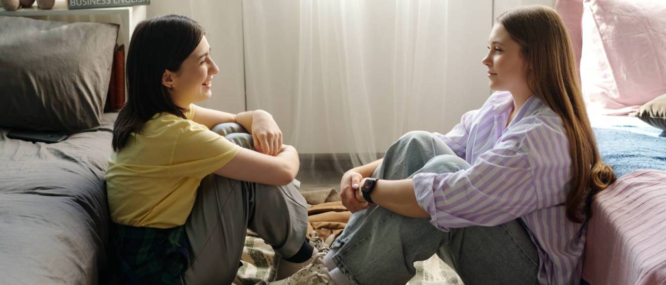 Two young women sitting on the floor between beds facing each other and talking in a bedroom, both looking relaxed and engaged in conversation with natural daylight streaming in, discussing a co-sign agreement.