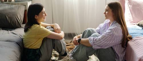 Two young women sitting on the floor between beds facing each other and talking in a bedroom, both looking relaxed and engaged in conversation with natural daylight streaming in, discussing a co-sign agreement.