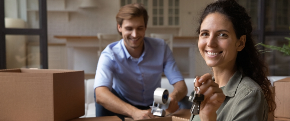 Happy couple with moving boxes and new keys, having compared the cost of living before choosing their new apartment.