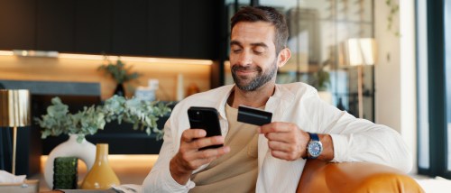 A smiling man sitting on a couch holds a smartphone and a payment card, about to pay a holding deposit online.