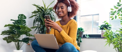 Woman wearing headphones and using her smartphone with a laptop on her lap, enjoying digital connectivity at home with RentCafe Internet.