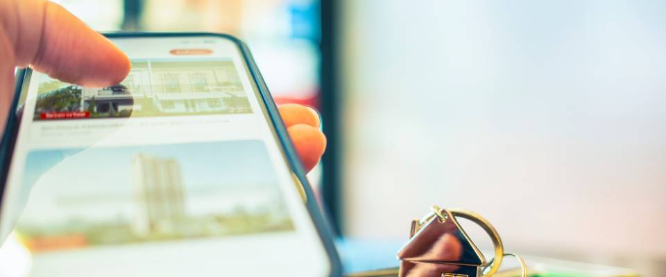 A close-up of a hand using a smartphone to search for real estate listings on screen, with a house key resting on the table, representing the process of finding an apartment for a new job.