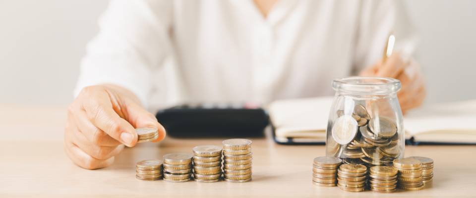 Close-up of a hand putting a coin into a clear glass jar filled with change on a bright desk in a minimal home scene representing personal savings, budgeting, money management, and future goals for apartments in Palo Alto.