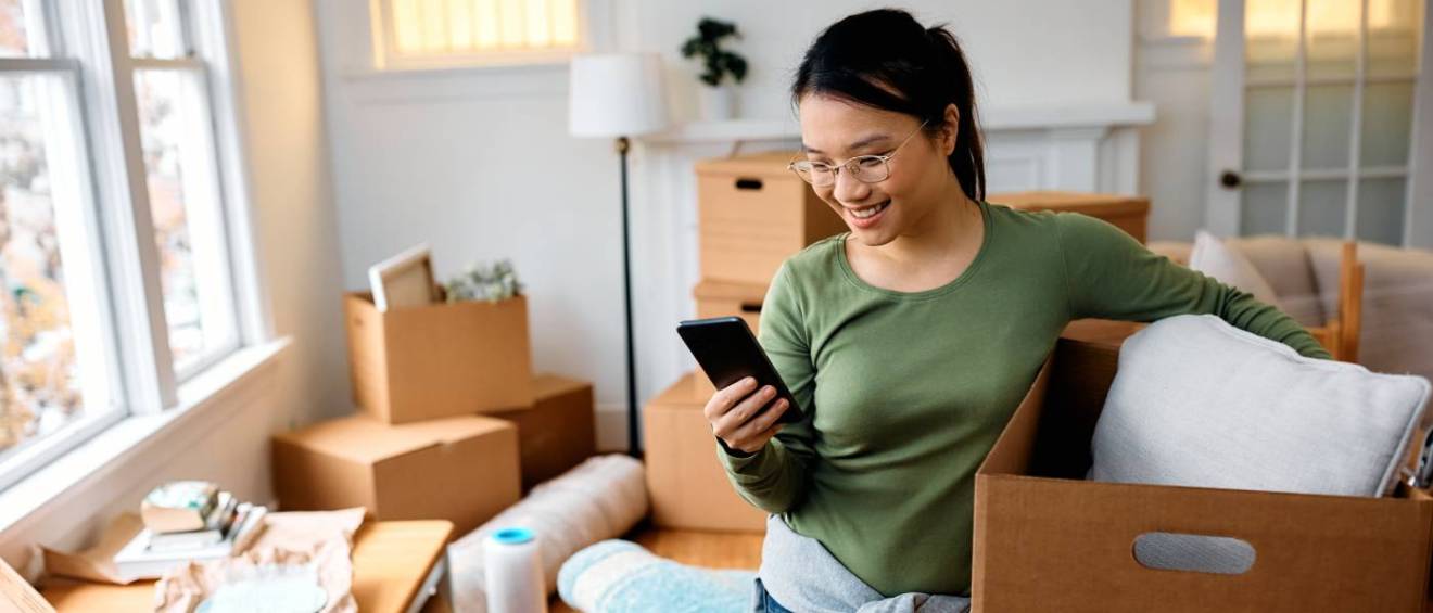 A young, happy woman smiles while using her smartphone as she unpacks her belongings in a new home, settling in after completing the first apartment rental process.