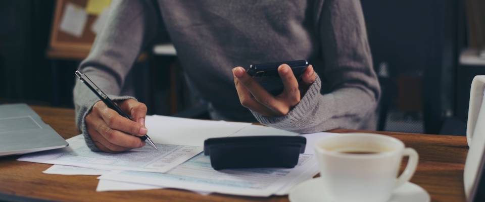 A woman wearing glasses uses her mobile phone and reviews financial documents, calculating expenses and budgeting as part of the first apartment rental process.