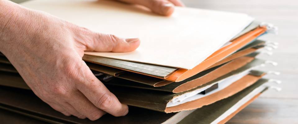 A close-up of hands grabbing file folders from a stack on a desk, with visible documents and paperwork, representing the organizational tasks involved in finding an apartment for a new job.