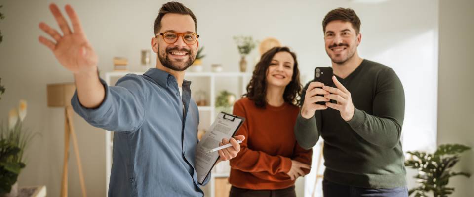 A real estate agent showing the interior of an apartment to a young couple, with the man recording the tour on his mobile phone as they navigate the first apartment rental process.